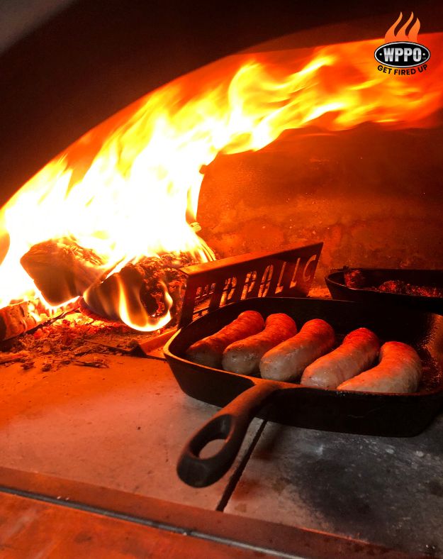 Sausages cooking in a cast iron skillet inside the WPPO Karma 55, with the wood fire burning alongside on the deck